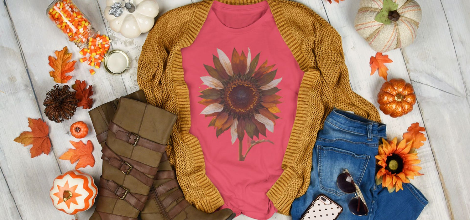 Pink shirt with sunflower design, mustard cardigan, blue jeans, and brown boots on a wooden surface with autumn decorations.